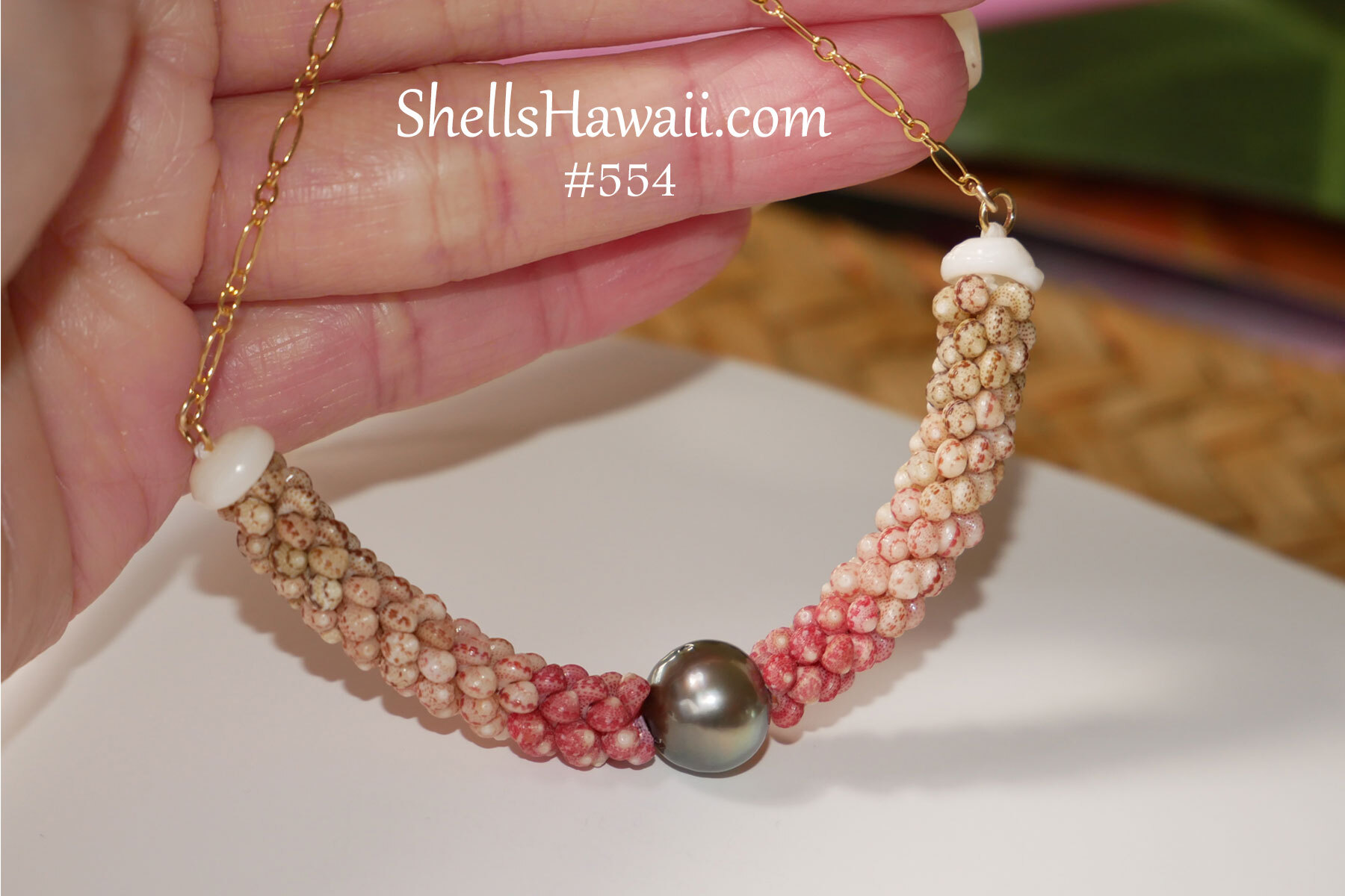 Close-up photograph of handcrafted Niʻihau shell jewelry necklace featuring Triple Poepoe style weaving created with genuine Kahelelani shells from Niʻihau. The necklace displays a soft natural ombr&eacute; color transition with Keʻokeʻo white, Mākuʻe brown, and ʻĀkala pink tones surrounding a dark Tahitian pearl centerpiece. Finished with a delicate 14k gold filled chain and white Momi shell accents.