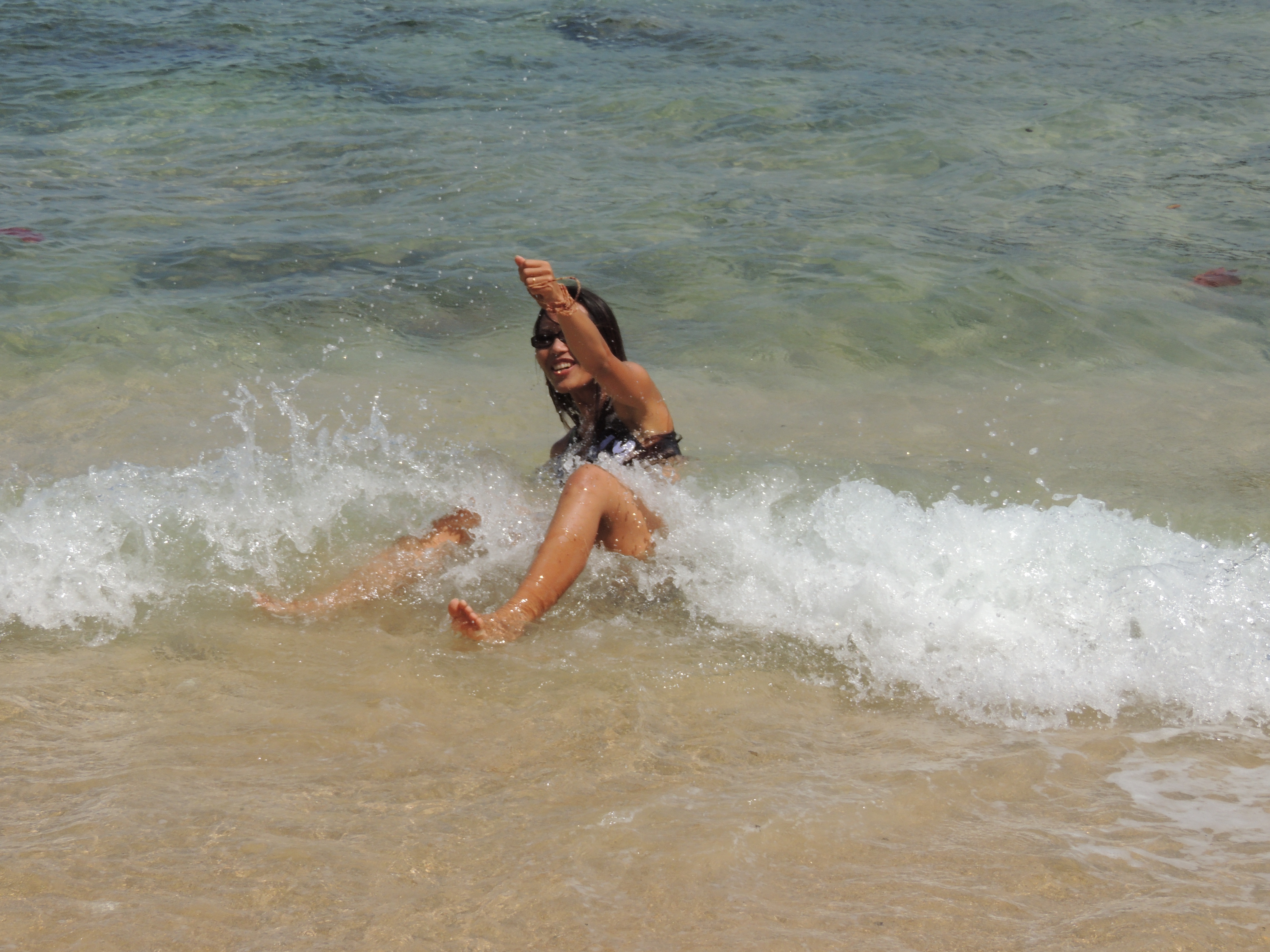 A playful behind-the-scenes moment of Janjira being knocked down by a wave during a Hawaiian jewelry photoshoot on Kauaʻi.