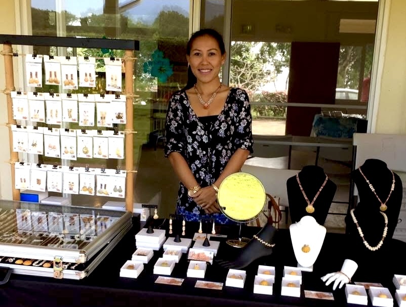 Meet the Maker – Janjira from ShellsHawaii ShellsHawaii artist Janjira standing behind her jewelry display table, featuring authentic Niʻihau shell earrings, necklaces, and leis. Each piece is handcrafted with aloha using natural shells from Niʻihau and Kauaʻi.