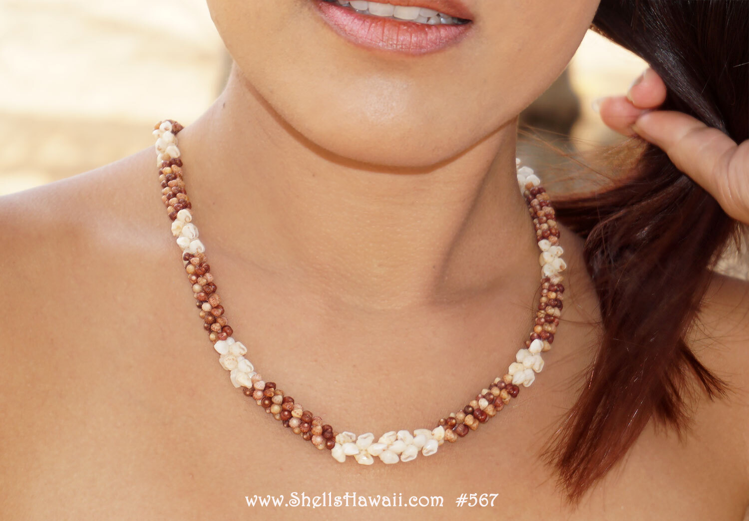 lose-up of handcrafted Hawaiian Kipona style shell lei featuring natural off white Momi shells combined with tiny burgundy and natural brown Kahelelani shells from Kauaʻi in an elegant traditional Hawaiian shell jewelry design displayed on Janjira&rsquo;s neck to show how the lei naturally lays when worn, with a soft beach scene background from Kauaʻi island.