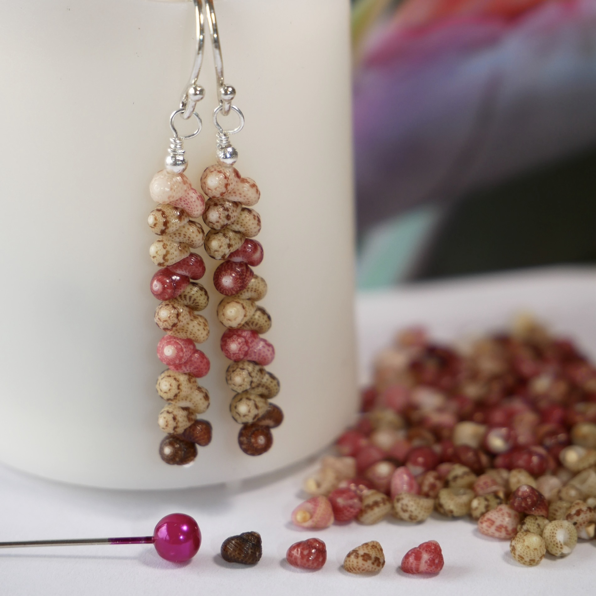Handcrafted Niʻihau shell earrings featuring genuine Kahelelani shells in green, brown, pink, and keʻokeʻo tones arranged in an organic natural minimalist design with sterling silver wire. The photo shows the dangling Hawaiian shell earrings displayed beside loose Kahelelani shells to help show the tiny shell sizes next to a regular size pink head pin.