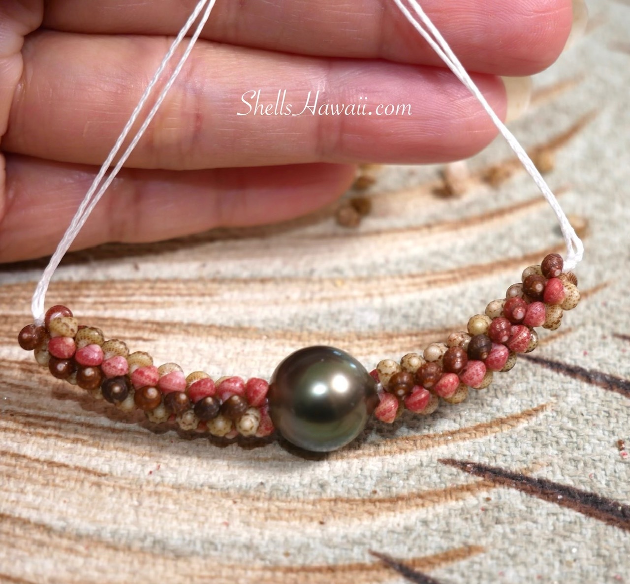 Colorful Niʻihau Kahelelani shells being woven around a Tahitian pearl to create the poepoe center section of a traditional Kipona style shell lei.