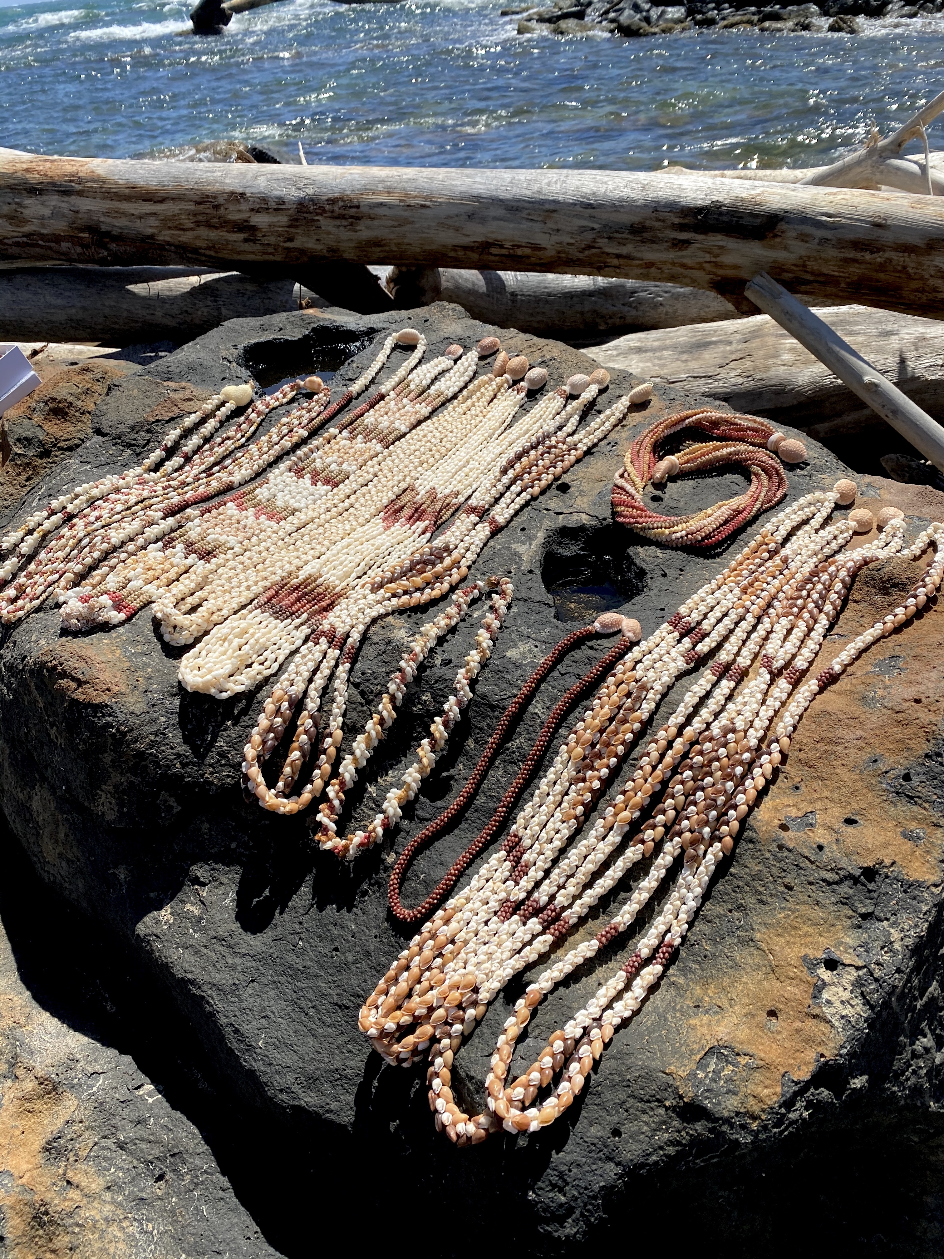 Traditional Hawaiian shell lei collection displayed on lava rocks near the ocean featuring genuine Poleho, Momi, and Kahelelani shells in authentic handcrafted Hawaiian jewelry styles.